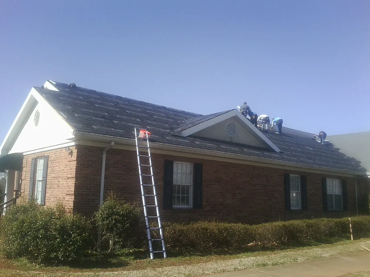 Crew tearing shingles off a brick ranch roof with a ladder set — Emergency Roof Repair work by our crew in Geneva